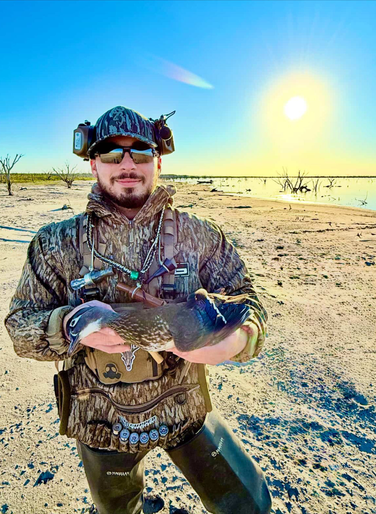 Man in NSG camouflage gear holding a bird in a desert landscape with a clear blue sky.