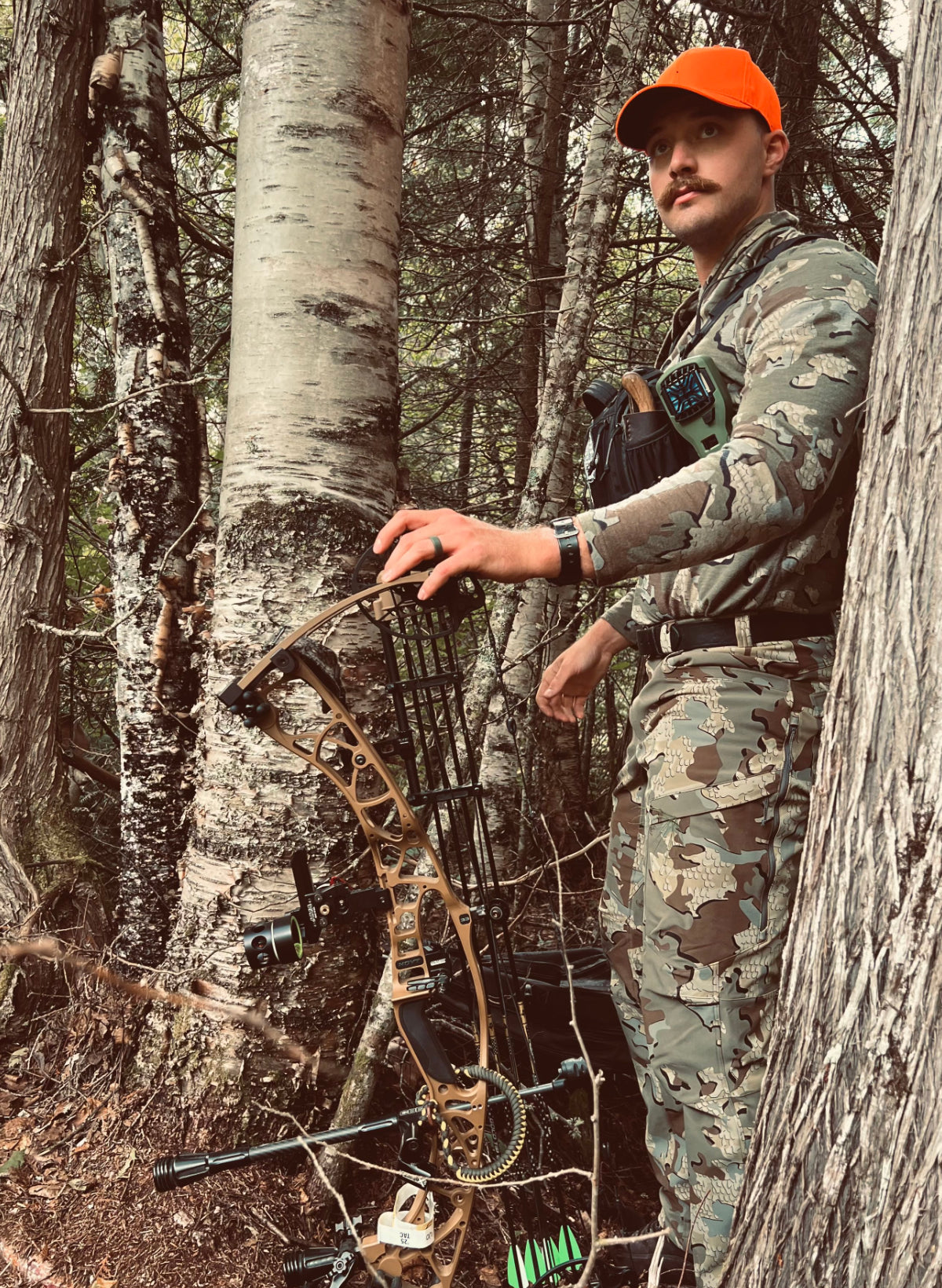 Man in camouflage clothing and orange cap with a bow in a forest setting